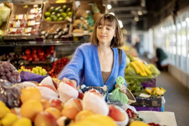 Genç, neşeli bir kadın marketten taze meyve seçiyor, yeniden kullanılabilir ağ torbasıyla alışveriş yapıyor. Sürdürülebilirlik ve organik yerel gıda konsepti