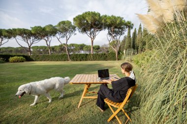 Genç, beyaz bir kadın dizüstü bilgisayardan bir şey izliyor ve yeşil Akdeniz çimlerinde, ahşap masada otururken çay içiyor. Hafta sonu, dinlenme ve uzak çalışma kavramı