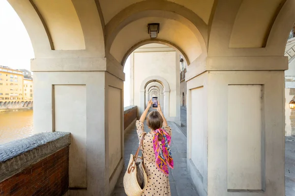 Young woman photographing on phone beautiful archway, visiting famous italian city Florence. Female tourist enjoys italian old architecture. Woman dressed in Italian style with colorful scarf in hair