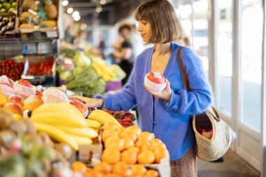 Kadın marketteki tezgahtan mango alırken, aynı zamanda tekrar kullanılabilir ağ torbasıyla meyve alışverişi yapıyor. Sürdürülebilirlik ve organik gıda konsepti