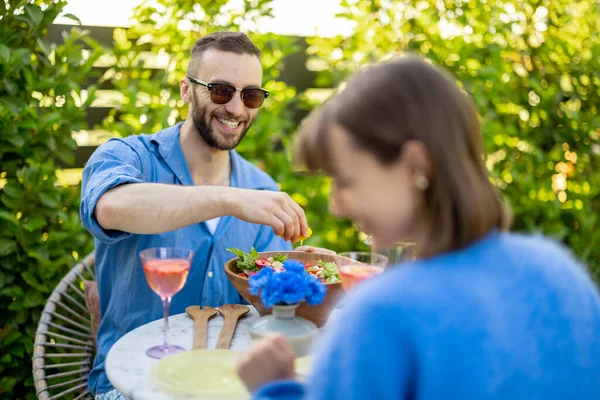裏庭で女性と昼食をとりながら 男はサラダにレモンを絞る 若い家族は屋外で健康的な食べ物を食べ 友人は庭で夏の時間を過ごす — ストック写真