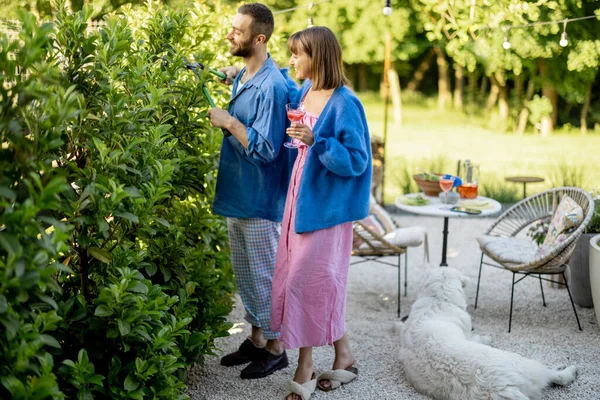 这对可爱的夫妇在他们乡间房子的后院里度过了夏天 男人修剪绿色的灌木丛 女人则喝一杯 与丈夫交谈 — 图库照片