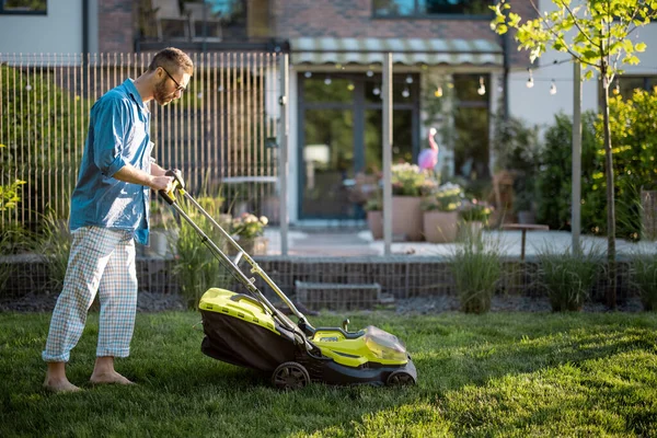 Man mows the lawn with lawn mower at backyard of his house. Husband takes care of garden on summer evening. Modern electric wireless garden equipment