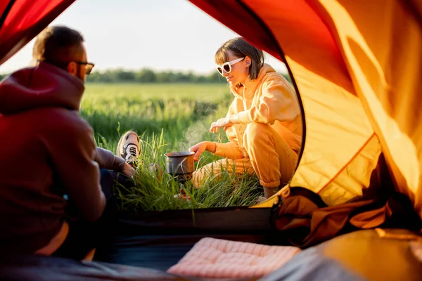 若いカップルは自然の中でテントと一緒に旅行中に食べ物を準備します 男と女はキャンプ場で夏を過ごす テントのドアからの眺め — ストック写真