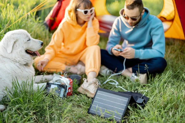 男人和女人在野外旅行时 与狗一起坐在营地里 带便携式太阳能电池板的充电器 现代旅行技术的概念 — 图库照片