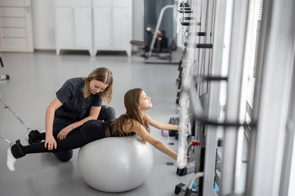 Rehabilitation specialist helping little girl to do exercises with fitness ball and decompression simulators at gym. Concept of physical therapy for back health and posture for kids