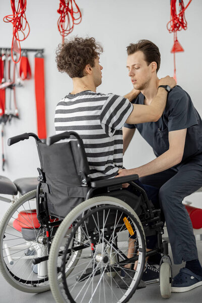 Rehabilitation specialist helps man to get up from a wheelchair at clinic. Concept of medical care for people with disabilities and physical rehabilitation