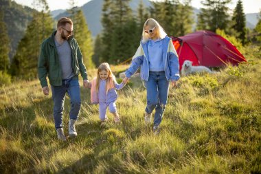 Genç beyaz bir aile ve küçük bir kız birlikte yeşil çayırlarda yürürken dağlarda çadır kurmuşlar. Kamp alanında mutlu aile tatilleri.
