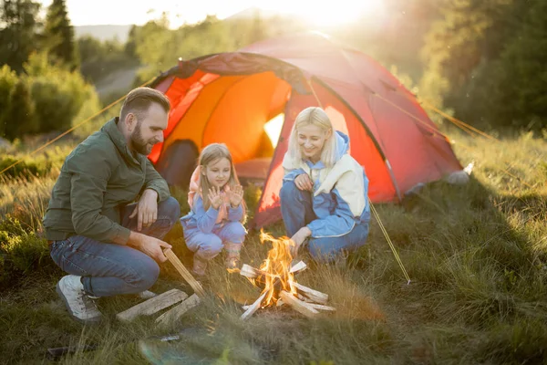 Young adult couple sit with a little girl by the campfire at campsite, traveling with tent on nature. Family spend summer time hiking in the mountains