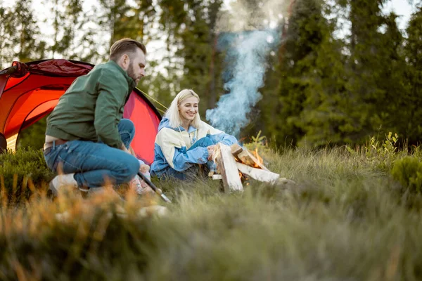 Young adult couple sit by the campfire, traveling with tent on nature. Family spend summer time hiking in the mountains