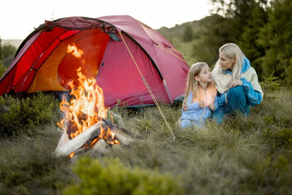 Young woman with her little girl sit by the campfire, bonding together while travel on nature. Mother with daughter warming up at campsite