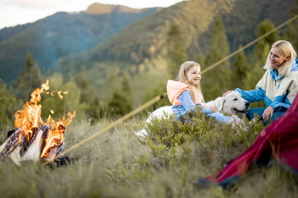 年轻的女人带着她的小女孩和小狗坐在篝火边 在大自然中旅行时紧紧地粘在一起 带着女儿的母亲在营地照看宠物 — 图库照片