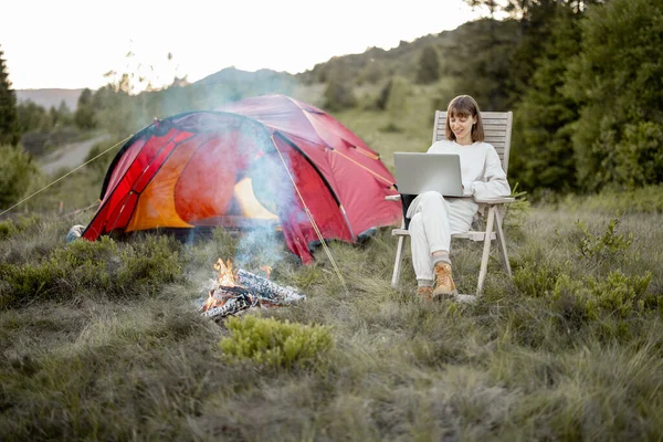 Young woman works on laptop while sitting relaxed on chair by the campfire, traveling with tent in the mountains. Concept of remote work and escape to nature