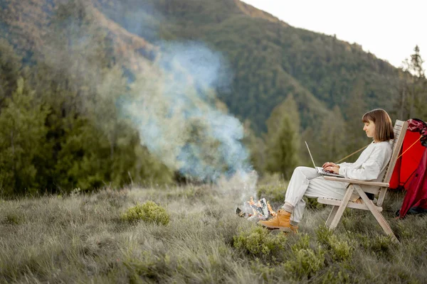 Young woman works on laptop while sitting relaxed on chair by the campfire, traveling with tent in the mountains. Concept of remote work and escape to nature