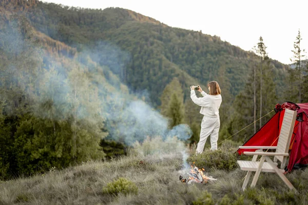 年轻女子站在篝火边 头戴帐篷 给美丽的山脉拍照 — 图库照片
