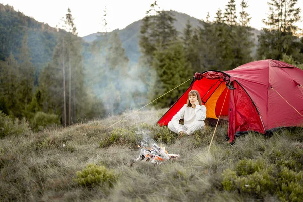 Young woman enjoys nature while sitting at campfire near camping tent in the mountains. Concept of escaping to nature