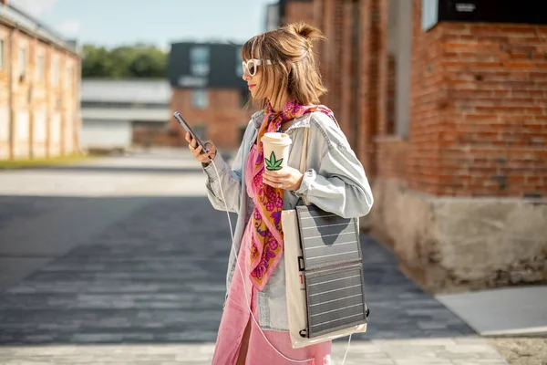 Young stylish woman walks with coffee and smart phone charging it from portable solar panel hanging on bag. Concept of modern and sustainable lifestyle