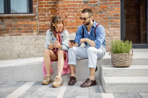 年轻时髦的男人和女人坐在一起 手持智能手机 在办公室附近与人交谈 咖啡休息时间的现代嬉皮士 — 图库照片