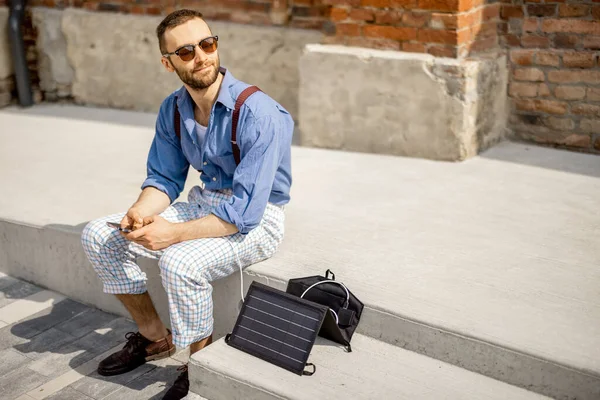 时尚男子使用智能手机 并从便携式太阳能电池板充电 而坐在街上室外 现代可持续生活方式和可再生能源 — 图库照片