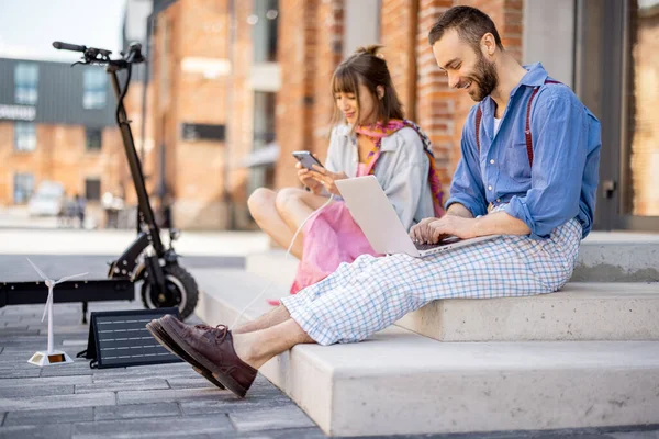 Stylish colleagues work on phone and laptop computer, charging them from portable solar panel, while sitting with electric scooter on a street at office district. Concept of sustainable lifestyle
