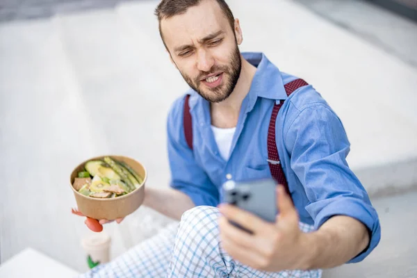 Stylish guy takes selfie or has online call on phone while eating healthy take away food during a lunch time outdoors. Concept of healthy vegan lifestyle on work