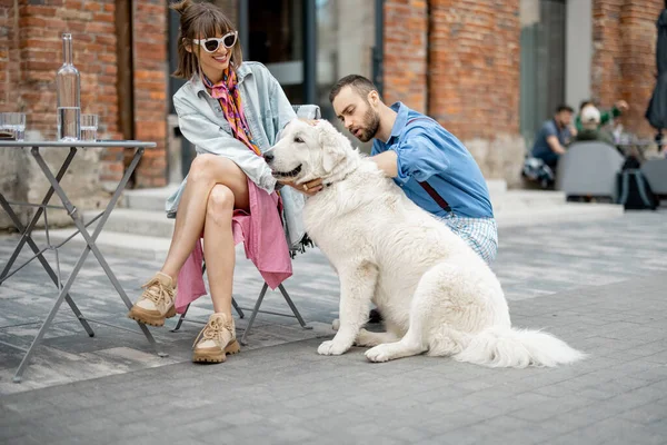 Stylish man and woman care adorable white dog while hanging out together at cafe terrace. Man flirting with woman caaring her dog