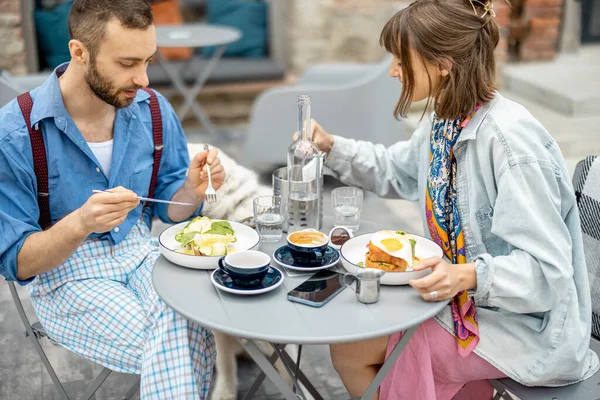 Stylish man and woman have delicious breakfast, eating and talking at cafe terrace. Friends spending time together during a breakfast