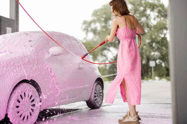 Young woman in pink dress washing her tiny car with nano foam at car wash during summer time. Easy and beautiful self-service at car wash concept