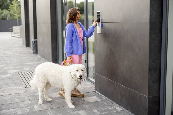 Молодая стильная женщина получает доступ к зданию, прикрепляя смартфон к домофону, стоя со своей собакой. Концепция современных технологий безопасности для доступа и интеллектуального дома