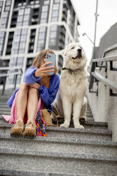 Cheerful woman takes selfie on phone with her dog while sitting on staircase at modern residential area outdoors. Spending leisure time and friendship with pets concept