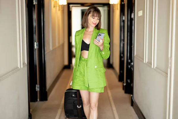 Young stylish business woman in green costume walks with suitcase and phone at the corridor of luxury hotel. Business travel and style concept
