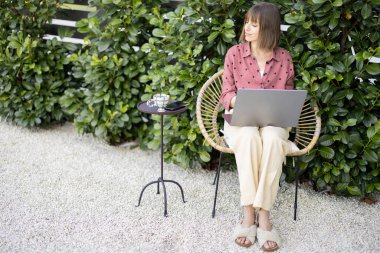 Genç kadın arka bahçedeki yeşil çalıların arka planında rahat rahat otururken dizüstü bilgisayarda çalışıyor. Rahat atmosferde uzaktan çalışma kavramı