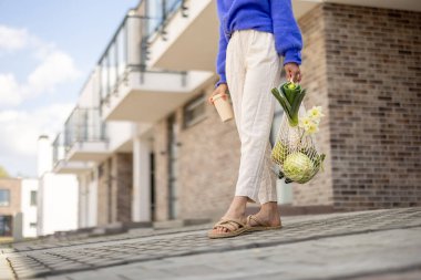 Kadının elinde bir çanta dolusu taze sebze ve yeniden kullanılabilir kahve fincanı var. Hem de yerleşim bölgesinde dikilirken. Sürdürülebilirlik kavramı, sağlıklı vejetaryen yiyecekler ve çevre dostu yaşam tarzı