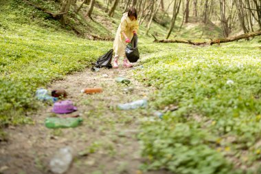Koruyucu elbiseli genç bir kadın ormanda dağınık plastik çöpleri topluyor. Yatak ekolojisi ve çevre kirliliği sorunu uzun zamandır çürümekte olan atıkla