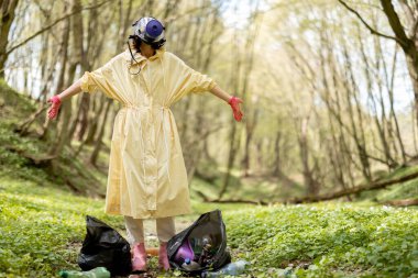 Gaz maskeli ve koruyucu elbiseli bir kadın elinde çöp torbalarıyla ormanda etrafa saçılmış plastik topluyor. Çevre kirliliği sorunu. Dünya konseptini koru.