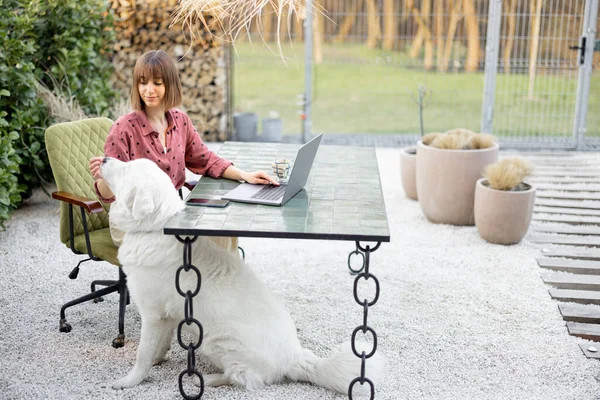 Young woman works on laptop and cares her dog while sitting at cozy outdoor workspace in the garden. Concept of remote work from comfortable home office or work during vacations