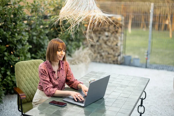 Young woman works on laptop at outdoor workspace in the garden. Concept of remote work from comfortable home office or work during vacations