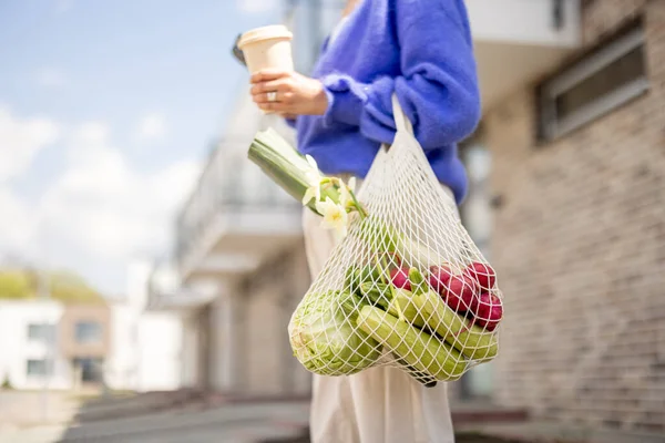 Woman holds mesh bag full of fresh vegetables and reusable coffee cup while standing at residential district, close-up. Concept of sustainability, healthy vegetarian food and eco-friendly lifestyle