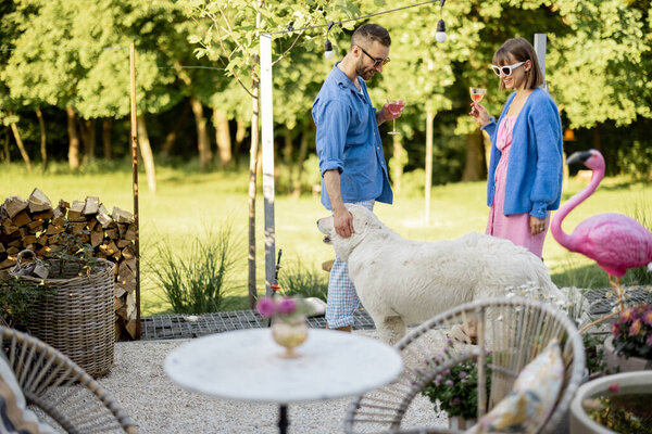 Young stylish couple hang out together and play with a dog at their beautiful backyard of country house. Man and woman spend summertime with pet outdoors