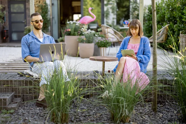 Young stylish couple work or spend leisure time with laptop computer and digital tablet on porch of their country house. Concept of remote work at cozy home atmosphere