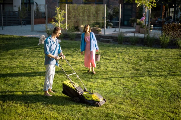 Man mows the lawn with lawn mower, spending summer time with his wife at backyard of country house. Young family take care of garden on summer evening