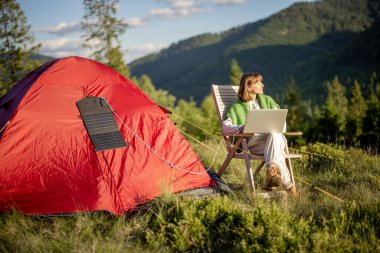 Genç kadın, dağlarda çadırla seyahat ederken dizüstü bilgisayarda çalışıyor. Çadırda asılı taşınabilir güneş paneli olan bir bilgisayar yükleniyor. Kamp alanında uzaktan çalışma ve yenilenebilir enerji kavramı
