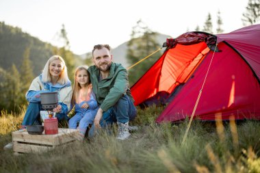 Portrait of a couple with little girl during picnic at campsite while traveling with tent in the mountains. Young family happily spending vacations on nature