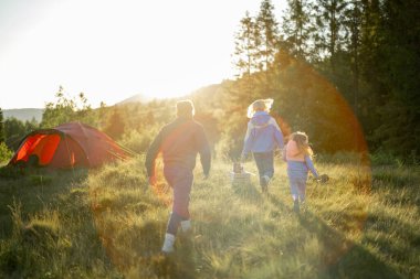 Young caucasian family with little girl walk together on green meadow while traveling with tent in the mountains during sunset. View from the backside. Image with lens flare