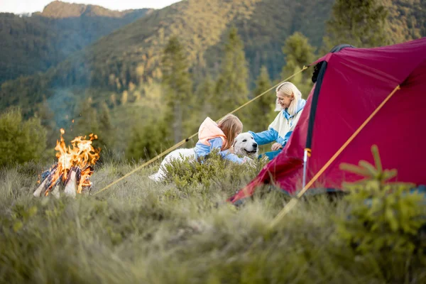 Young woman with her little girl and dog sit by the campfire, bonding together while travel on nature. Mother with daughter care pet at campsite