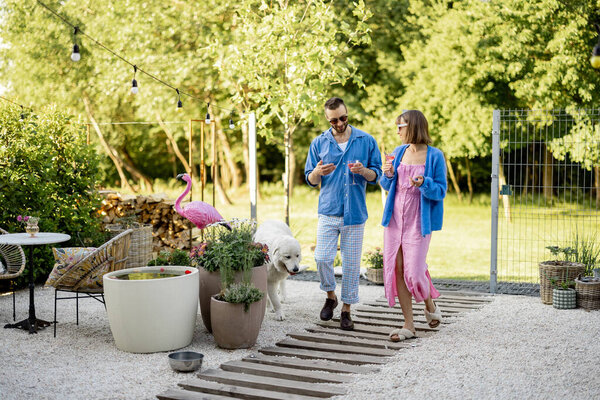 Young stylish couple have romantic conversation while walking with a drink at their beautiful backyard of country house. Young man and woman spend summer time happily