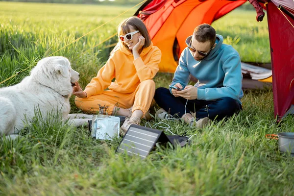 男人和女人在野外旅行时 与狗一起坐在营地里 带便携式太阳能电池板的充电器 现代旅行技术的概念 — 图库照片