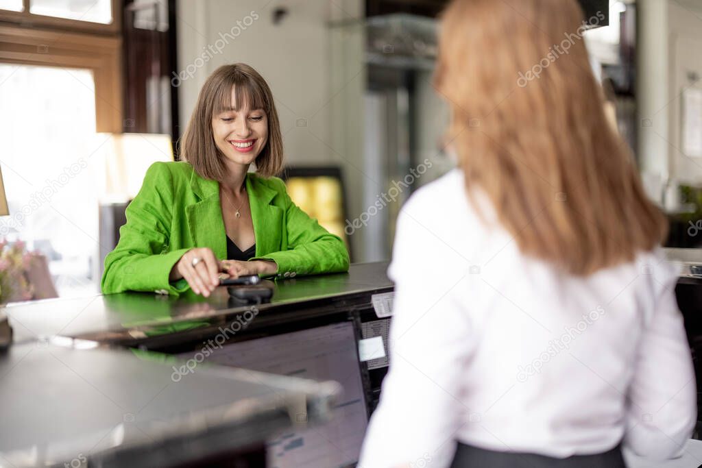 Young cheerful business woman pays contactlessly by phone at the hotel reception. Business trip and hospitality service concept