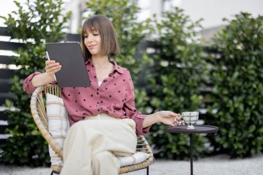 Genç yaratıcı kadın evinin arka bahçesinde kahve fincanıyla rahatça otururken dijital tablet üzerinde çalışıyor. Uzaktan çalışma kavramı ya da dışarıda aletlerle boş zaman geçirme kavramı