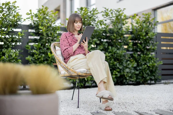 Young creative woman working on digital tablet while sitting relaxed on chair at backyard of her house. Concept of remote work or leisure time with gadgets outdoors